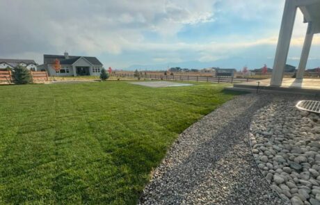 Large backyard with green grass, a gravel path, wooden fence, and a distant view of hills under a partly cloudy sky.