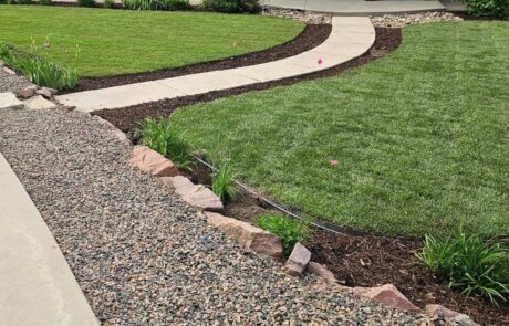 A neatly landscaped front yard with a curved pathway, green lawn, rocks, and mulch, leading to a modern gray house with large windows.