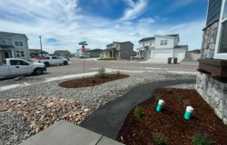 Suburban neighborhood with modern houses, a street with parked vehicles, a stop sign, and a landscaped yard with a stone path and mulch. Cloudy sky above.