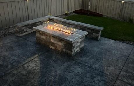 Stone fire pit with a rectangular flame in a backyard, surrounded by a stone bench on a stamped concrete patio. Fence and small tree in the background.