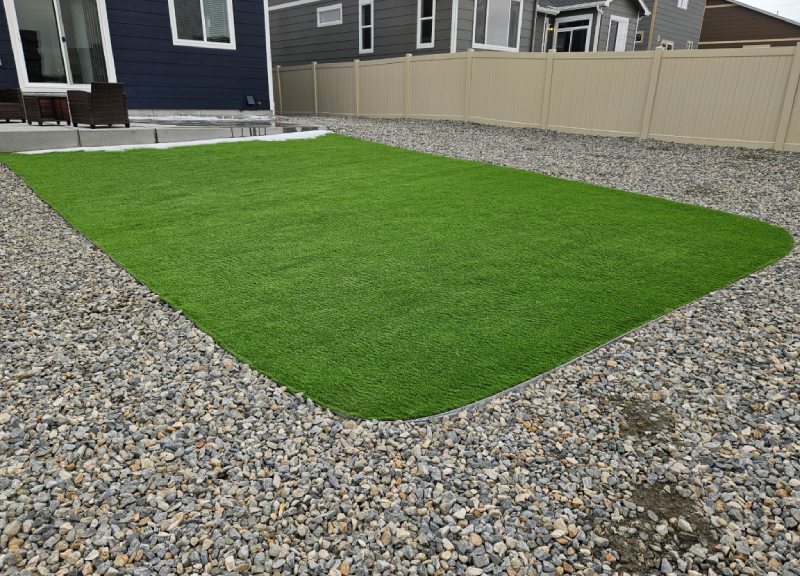 A backyard with a rectangular patch of artificial grass surrounded by gravel, enclosed by a tan fence and neighboring houses in the background.