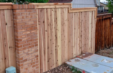 Wooden fence with a brick column on the left, next to utility boxes on the ground.