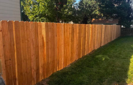 A long, wooden fence runs alongside a green lawn with trees and a house in the background under a clear sky.