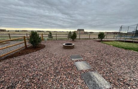 Backyard with circular stone fire pit, stepping stones leading to it, surrounded by a gravel area, wooden fence, some bushes, and a trampoline on the right side under a cloudy sky.