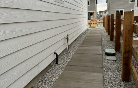 A narrow concrete walkway runs alongside a house with white siding, bordered by gravel and a wooden fence on the right. Other houses are visible in the background.