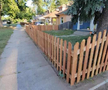 A wooden picket fence runs alongside a sidewalk, separating it from a residential front yard with trees and houses in the background.