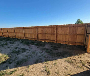 A wooden privacy fence with a gate borders a patch of dry, grassy ground under a clear blue sky.