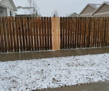 A wooden fence with two new planks replacing old ones, set beside houses in a snowy landscape.