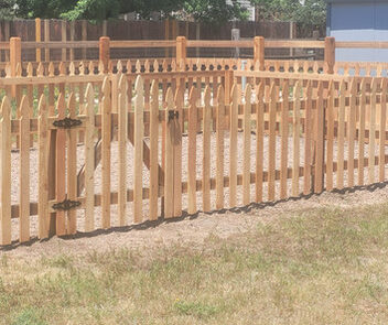 Wooden picket fence enclosing a garden area with a wooden gate in a grassy yard. Trees and a shed are in the background.