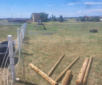 A grassy yard with a partially installed fence, wooden posts, and fencing materials scattered on the ground, set under a clear blue sky.