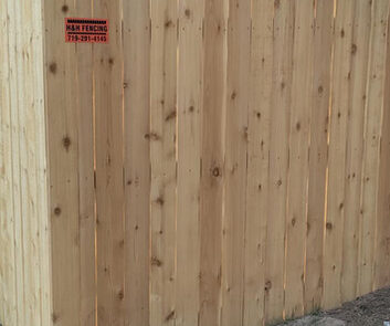 Wooden privacy fence with vertical planks. A sign on the fence reads "M & M Fencing 719-231-4345." Grass is visible at the bottom.