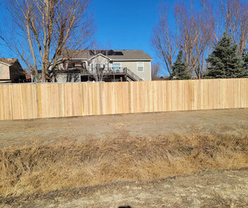 A wooden fence stands in front of a house with a clear blue sky and bare trees in the background. A person's shadow is visible in the foreground.