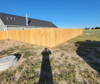 A long wooden fence surrounds a grassy yard next to a single-story house under a clear blue sky. A person's shadow is visible on the ground.