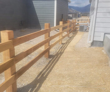 A wooden fence separates two houses with grey siding, casting shadows on a gravel and straw-covered ground.