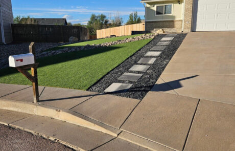 A house with a neatly trimmed lawn, stone pathway, and driveway leading to a garage. A white mailbox is positioned at the edge of the sidewalk.