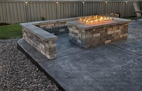 A stone fire pit with glass wind guard on a concrete patio, surrounded by a gravel perimeter and a wooden fence in the background.