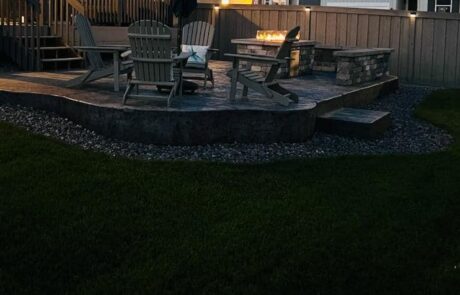 A patio with Adirondack chairs surrounds a lit fire pit. The area is elevated with stone steps leading down to a grassy lawn.