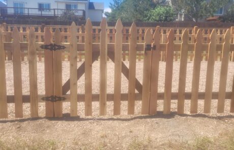 Wooden picket fence with a gate, featuring black hinges and a latch, encloses a gravel area. A house and trees are visible in the background.