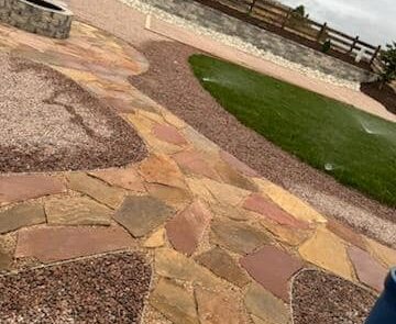 Backyard with stone pathways, grassy area, raised stone fire pit, and wooden fence under a cloudy sky.