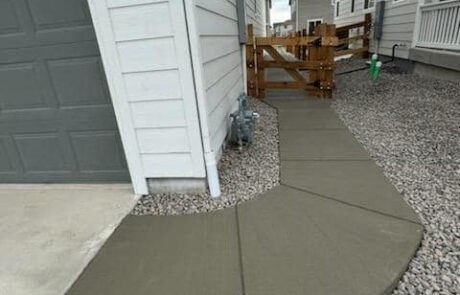 Concrete path curves around a gray garage, bordered by gravel and leading to a wooden gate between white houses.