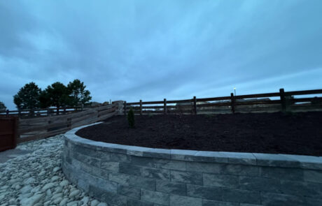 A stone wall with a bed of dark soil and a wooden fence on top under a cloudy, blue evening sky.