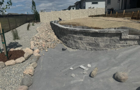 Backyard view with a partially constructed stone retaining wall, gravel path, scattered rocks, and a wooden fence under a cloudy sky.