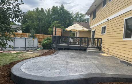 Backyard with a gray stamped concrete patio, above-ground pool, and deck with an umbrella. Large yellow house and trees in the background.